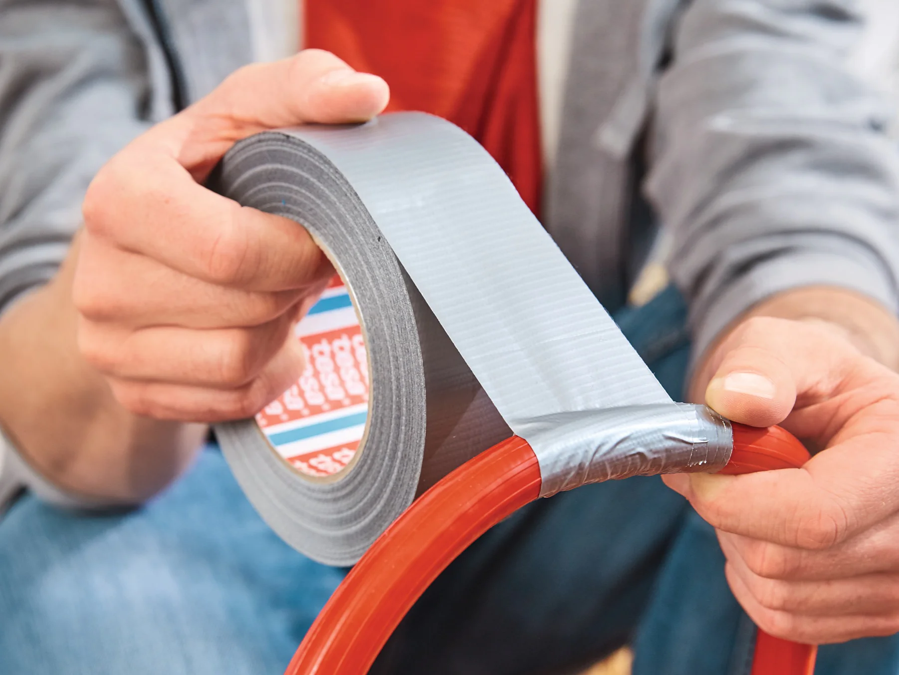 A person wearing a gray hoodie and blue jeans is using silver tesa tape to seal or repair a red hose. They are sitting and focusing on wrapping the tesa tape around the hose securely. (This text has been generated by AI)