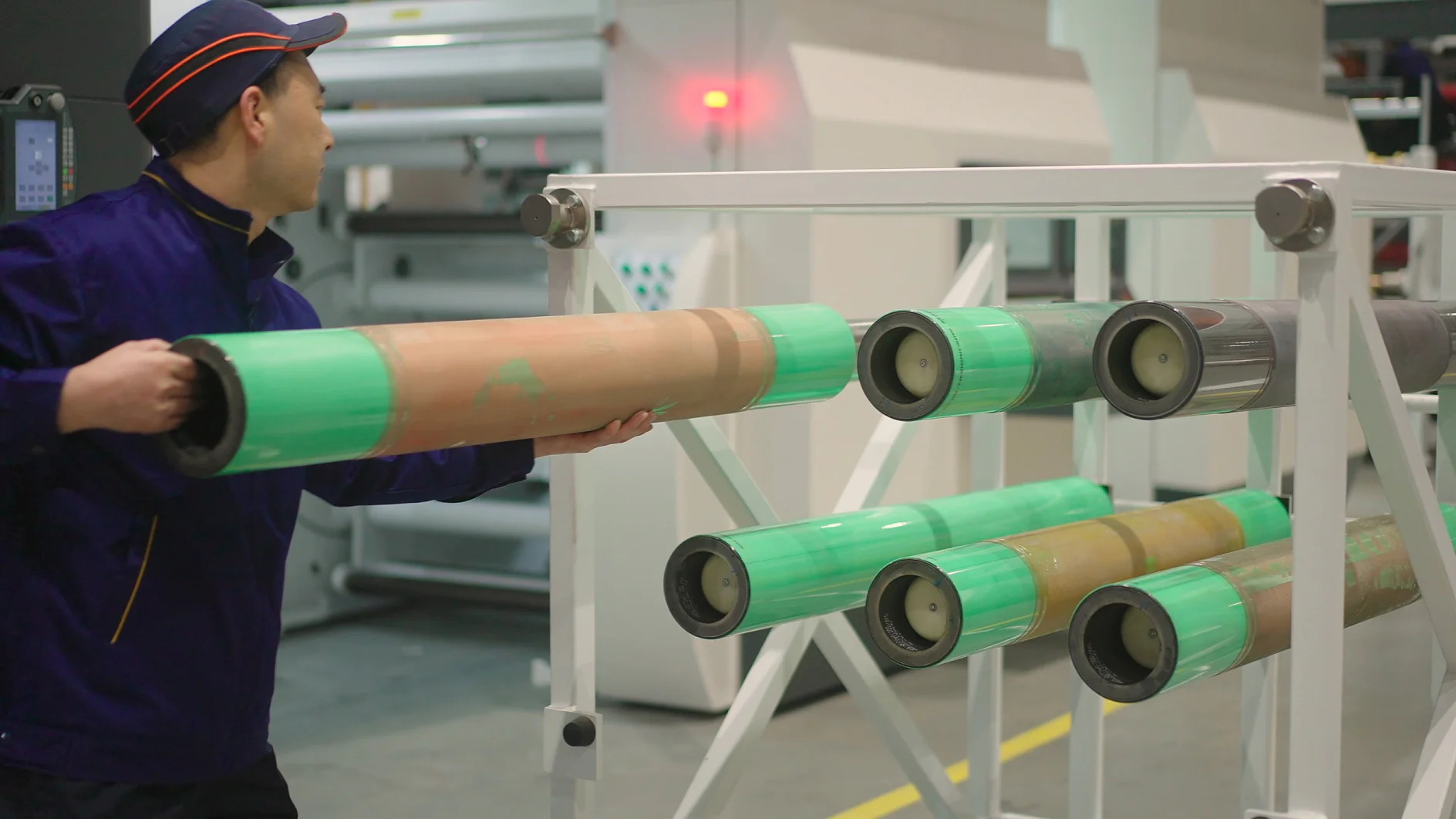 A person in a blue uniform operates machinery in a factory, handling a large cylindrical component wrapped in green and brown tesa tape. Other similar components are mounted on a white metal frame nearby. (This text has been generated by AI)