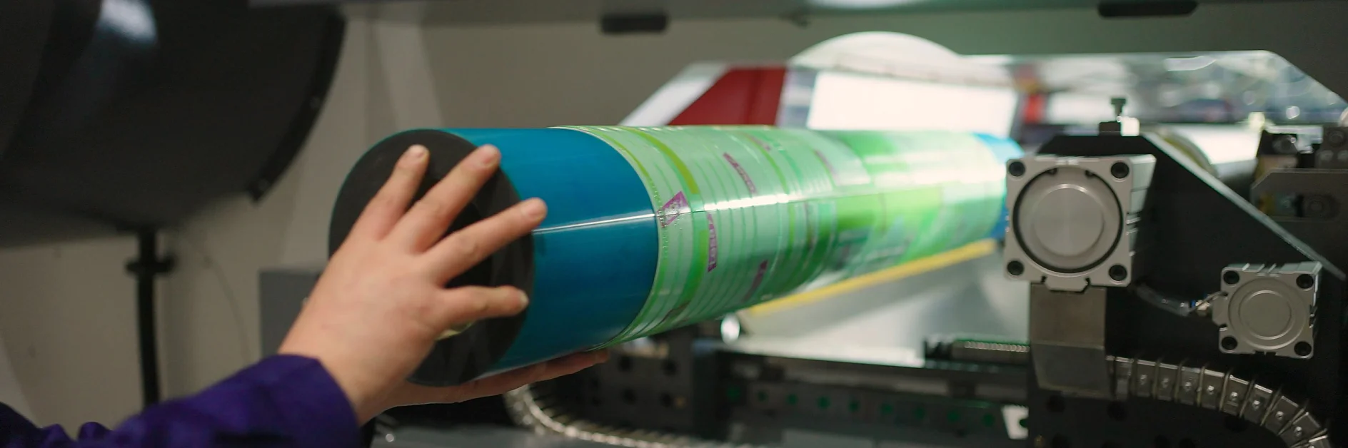 A person places a cylindrical green and blue printing plate into a large printing machine using tesa tape. The machine features various buttons and indicators. The setting seems to be in an industrial or manufacturing environment. (This text has been generated by AI)