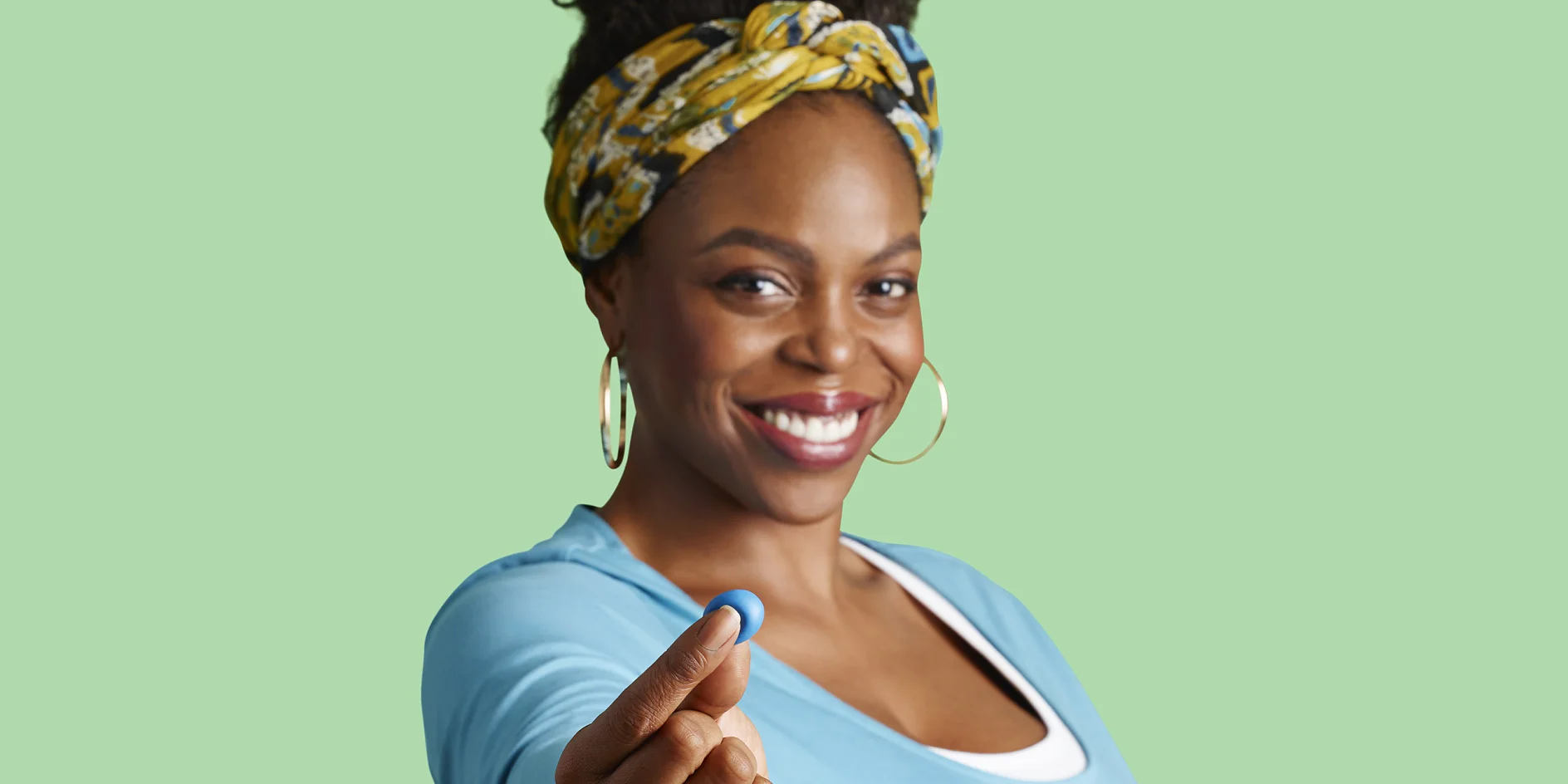 A woman in a blue top is smiling and holding a small blue object between her fingers. She has a multicolored headband and gold hoop earrings. The background is a solid light green color. (This text has been generated by AI)