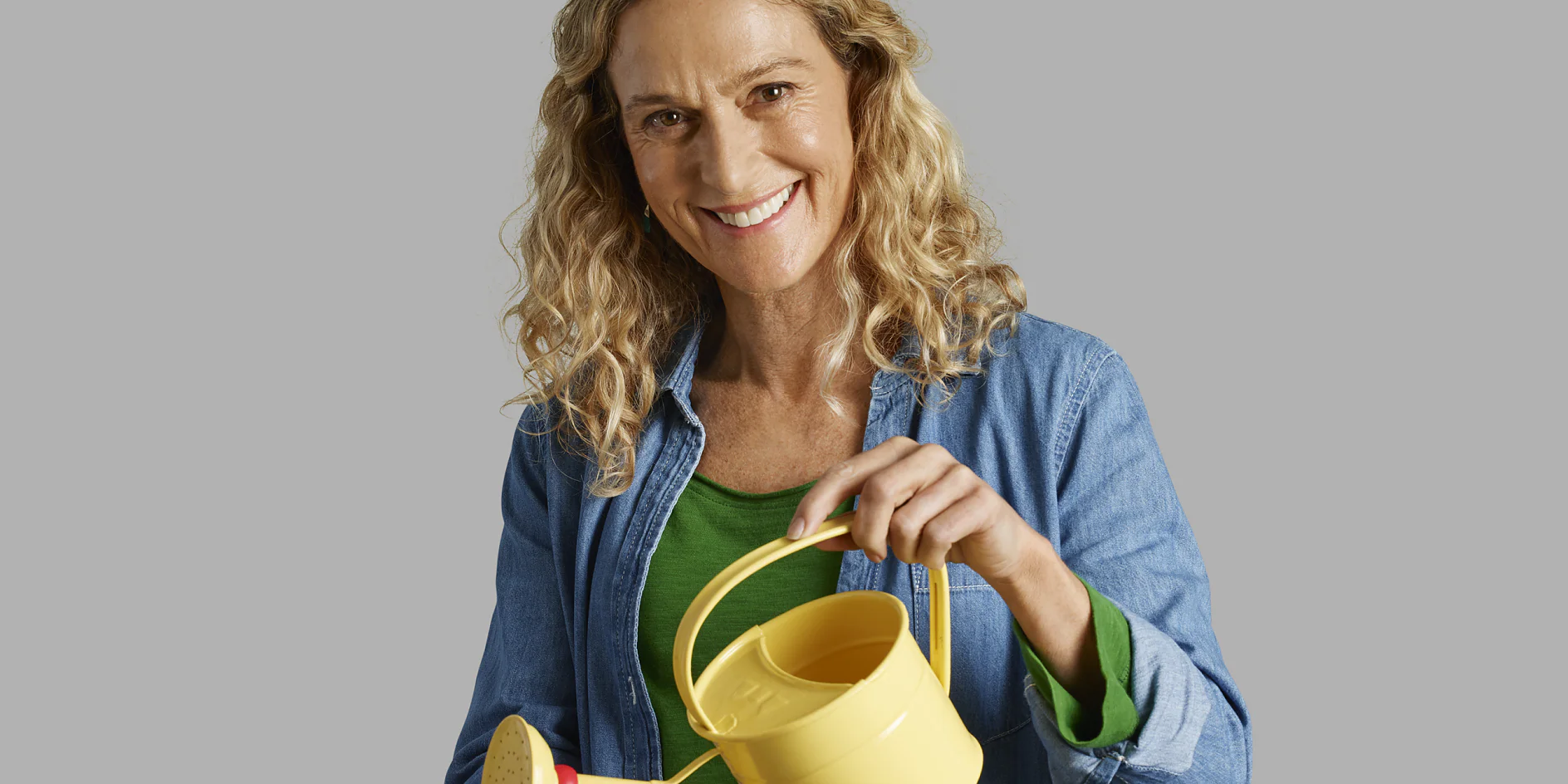 A person with curly hair is smiling and holding a yellow watering can. They are wearing a green shirt and a denim jacket. The background is plain gray. (This text has been generated by AI)