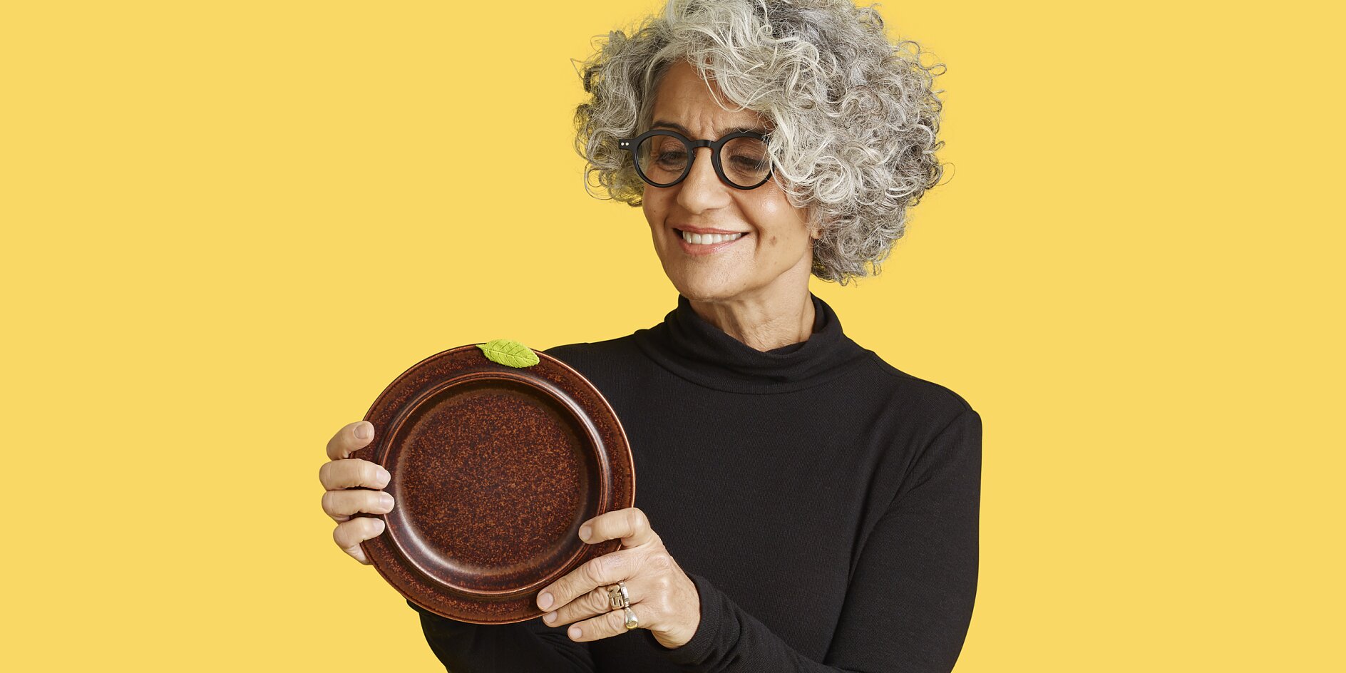 A person with curly gray hair and glasses is holding a brown plate with both hands, smiling. They wear a black top and stand against a solid yellow background. (This text has been generated by AI)