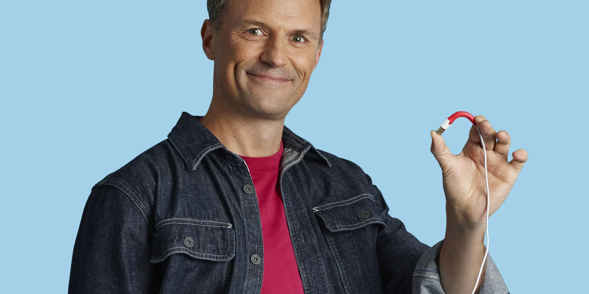 A person in a denim shirt and red t-shirt is holding a magnetic charging cable with a red end against a light blue background. The person is smiling and looking at the camera. (This text has been generated by AI)