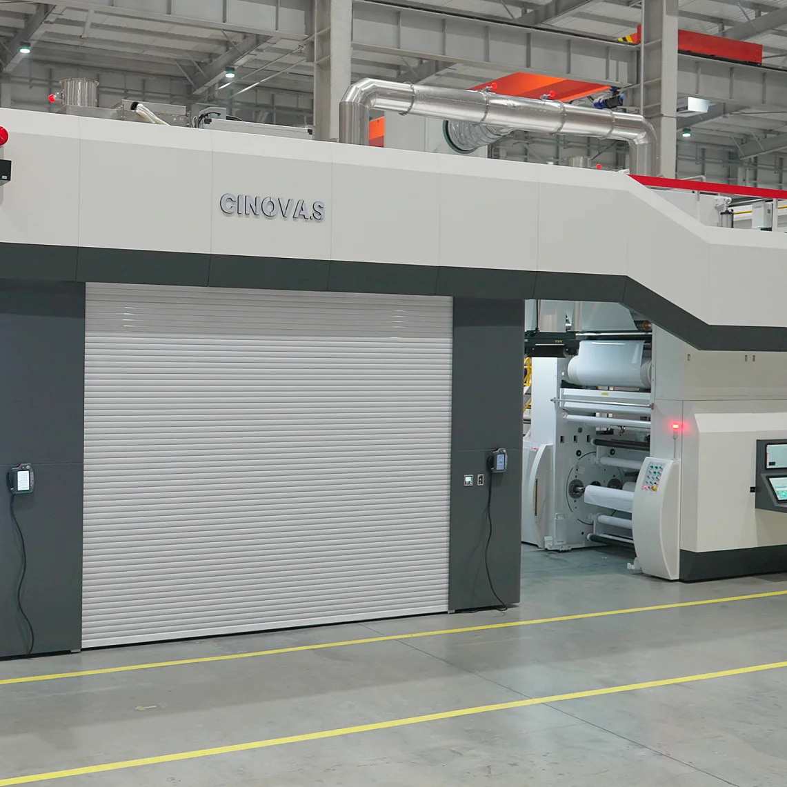 Large industrial printing machine in a factory setting. The machine is gray and white with control panels and display screens. The floor is concrete, and the area is well-lit with overhead lights. Yellow lines are marked on the floor for guidance using tesa tape. (This text has been generated by AI)