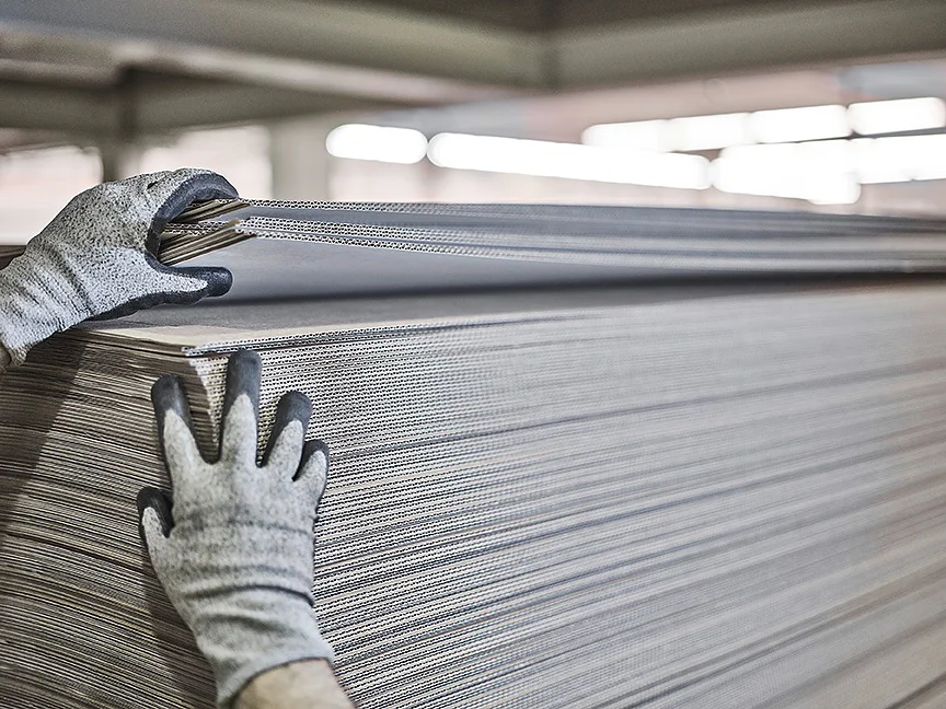 Hands wearing gloves are adjusting a large stack of corrugated cardboard sheets in a warehouse or industrial setting. The sheets are arranged in a neat pile, with strips of tesa tape securing the layers together. The background shows bright, blurred lighting. (This text has been generated by AI)