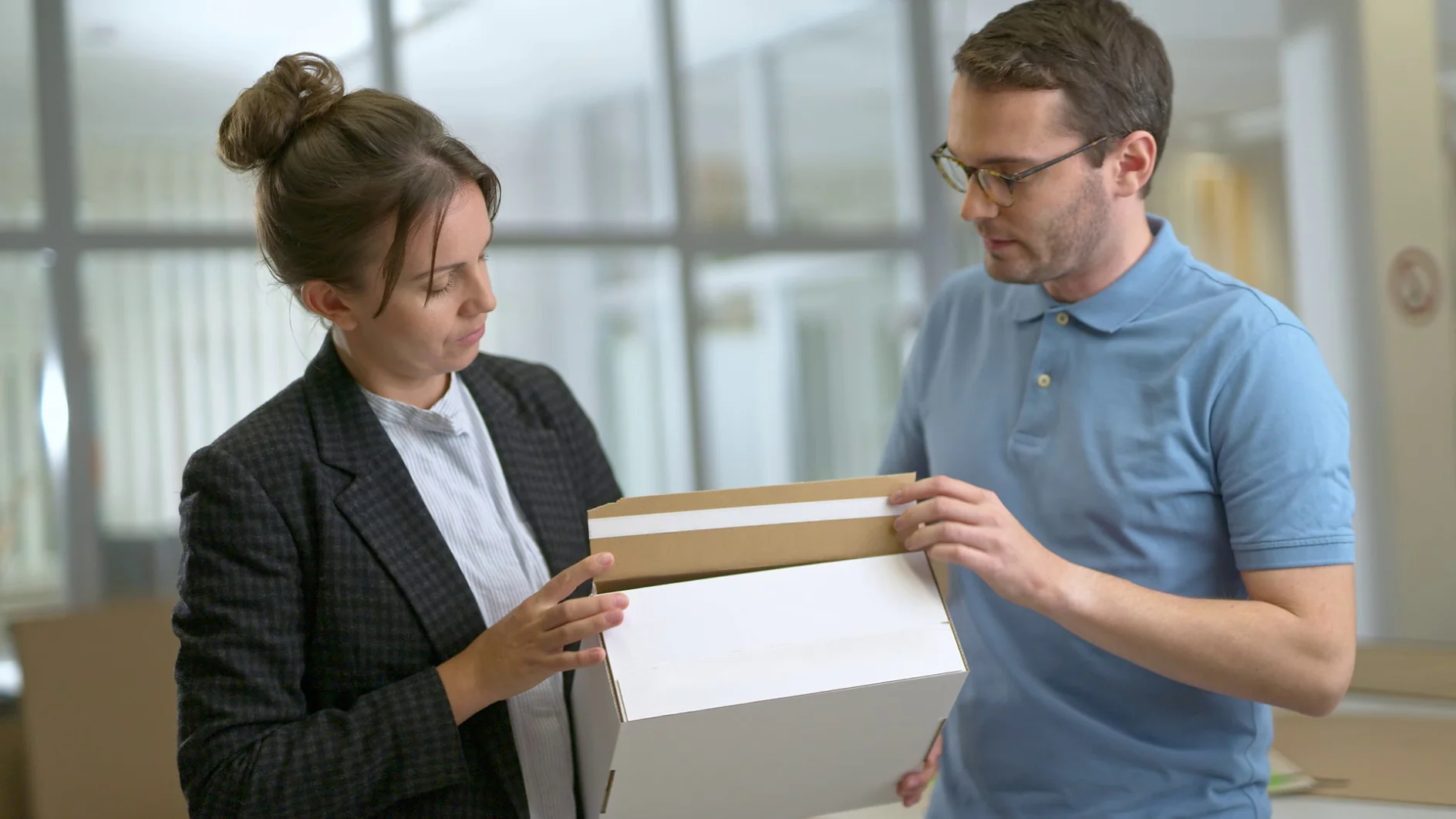 A man in a blue polo shirt and glasses is holding a white cardboard box secured with tesa tape, while a woman in a checkered blazer is looking at it. They are standing in an office setting with glass partitions in the background. (This text has been generated by AI)