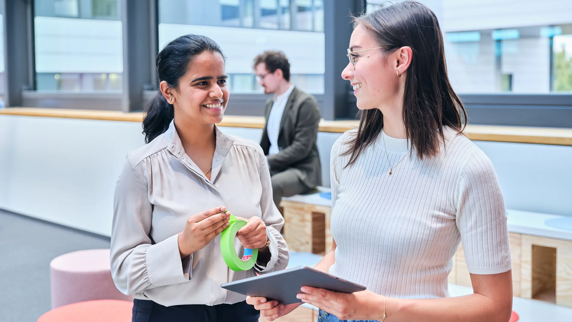 Two women are standing indoors having a conversation. One is holding a roll of green tesa tape, and the other is holding a tablet. A man is seated in the background. The room has large windows and modern furniture. (This text has been generated by AI)