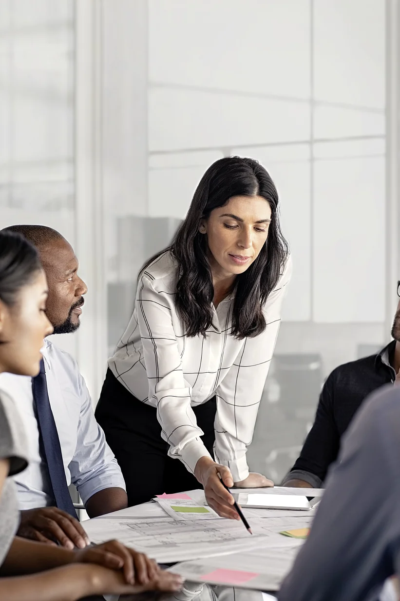 A woman stands and points at a document on a table during a meeting with four seated colleagues. They are in a modern office with glass walls. Papers and tesa tape sticky notes are spread across the table. (This text has been generated by AI)