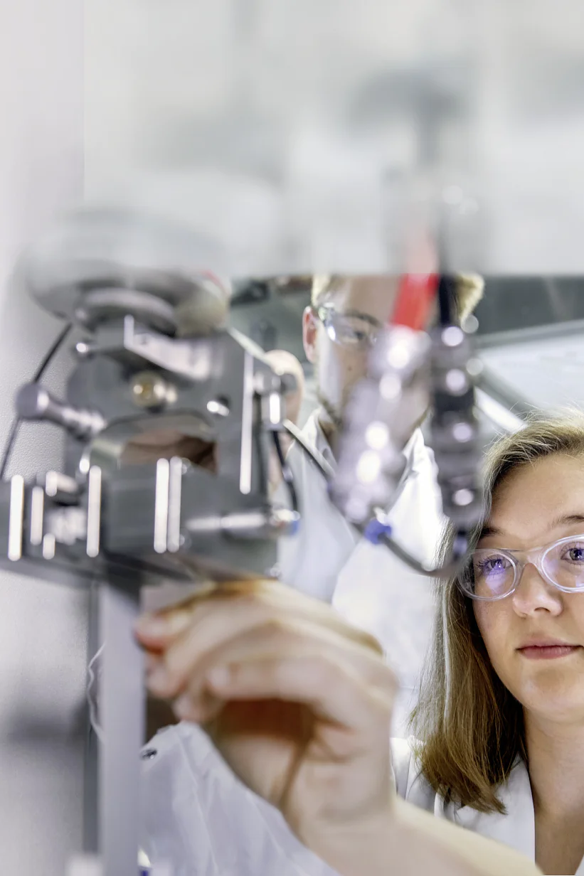A woman wearing safety goggles operates lab equipment. She adjusts a metallic component under a mechanical setup using tesa tape. The environment appears to be a laboratory with a focus on scientific research or experimentation. (This text has been generated by AI)