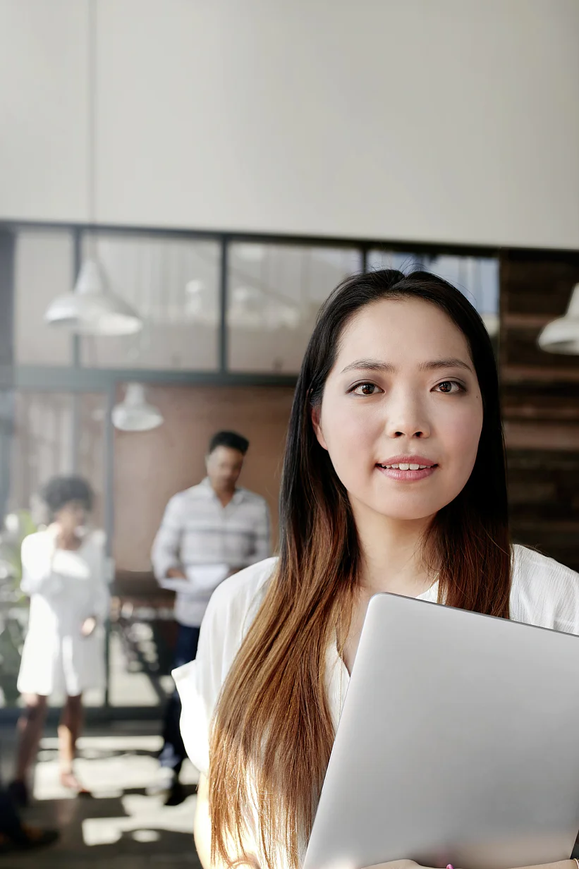 A woman holding a laptop stands in the foreground of a modern office. She looks directly at the viewer. In the background, three people engage in conversation near a glass-walled meeting room with pendant lights and tesa tape. (This text has been generated by AI)
