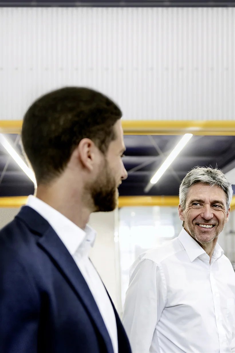 Two men in white shirts and blazers are engaged in conversation in an industrial setting with overhead lights. One is facing the camera smiling, while the other has his back to the camera. Yellow beams and rolls of tesa tape are visible in the background. (This text has been generated by AI)
