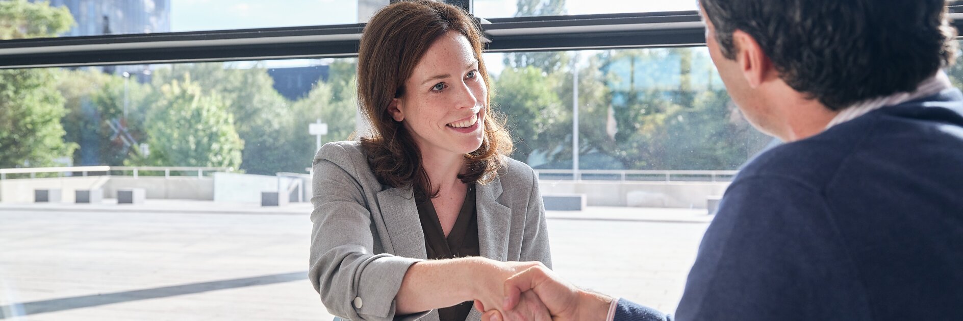 A man and a woman are sitting at a table in a brightly lit office with large windows. They are shaking hands across the table, with a notepad, pen, and roll of tesa tape placed in front of them. Trees and a glass building are visible outside. (This text has been generated by AI)