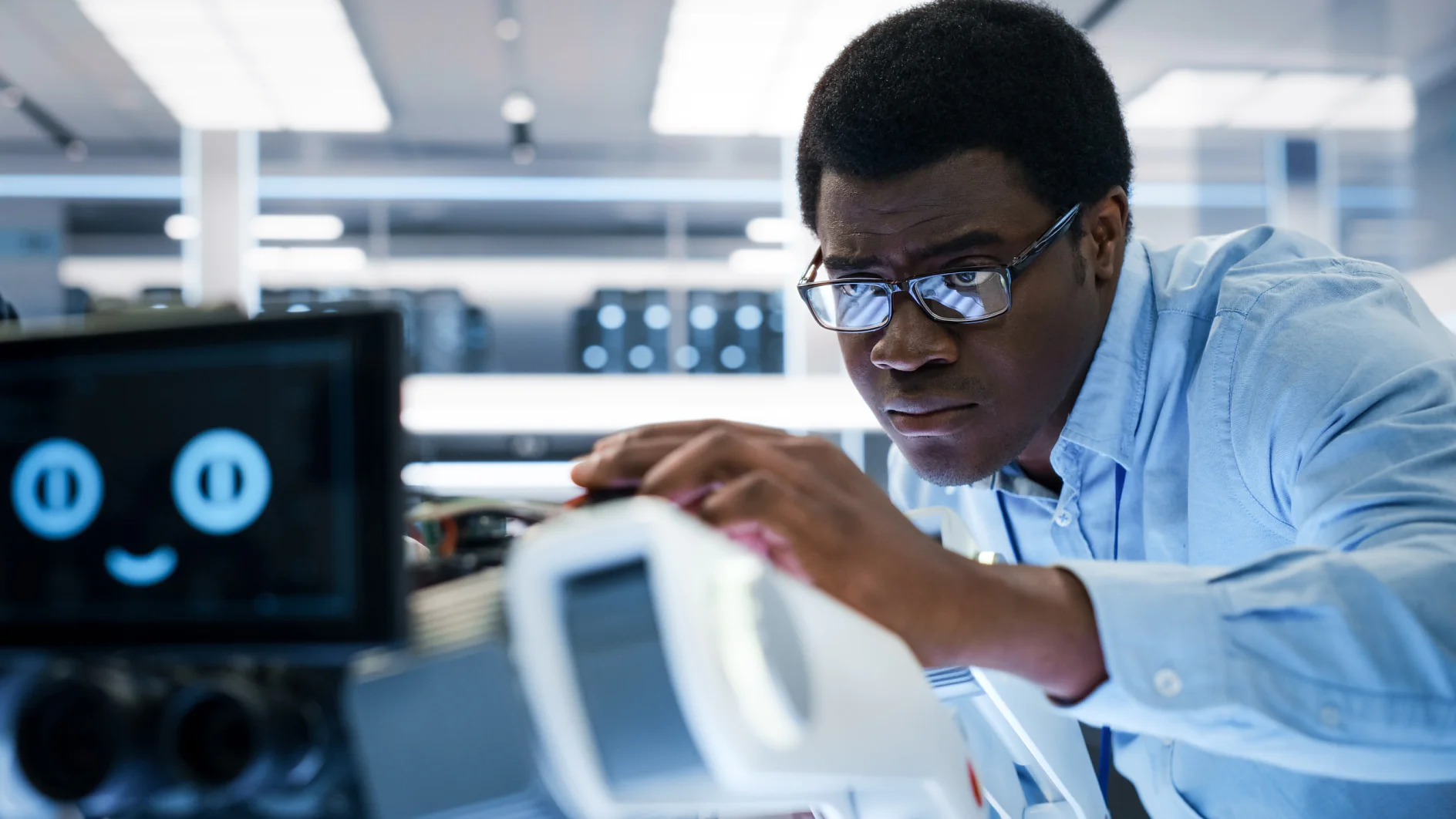 A person wearing glasses and a light blue shirt is adjusting a piece of equipment in a laboratory setting using tesa tape. A screen displaying a smiley face is visible in the foreground, while the background features blurred lab equipment. (This text has been generated by AI)