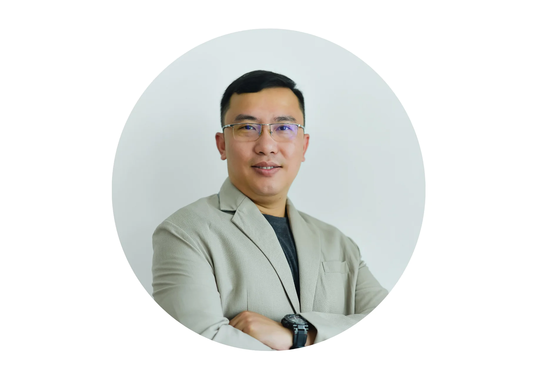 A man with short black hair and glasses is wearing a light beige blazer over a dark shirt. His arms are folded, and he is smiling slightly. The background is plain white, and the image is circular. (This text has been generated by AI)