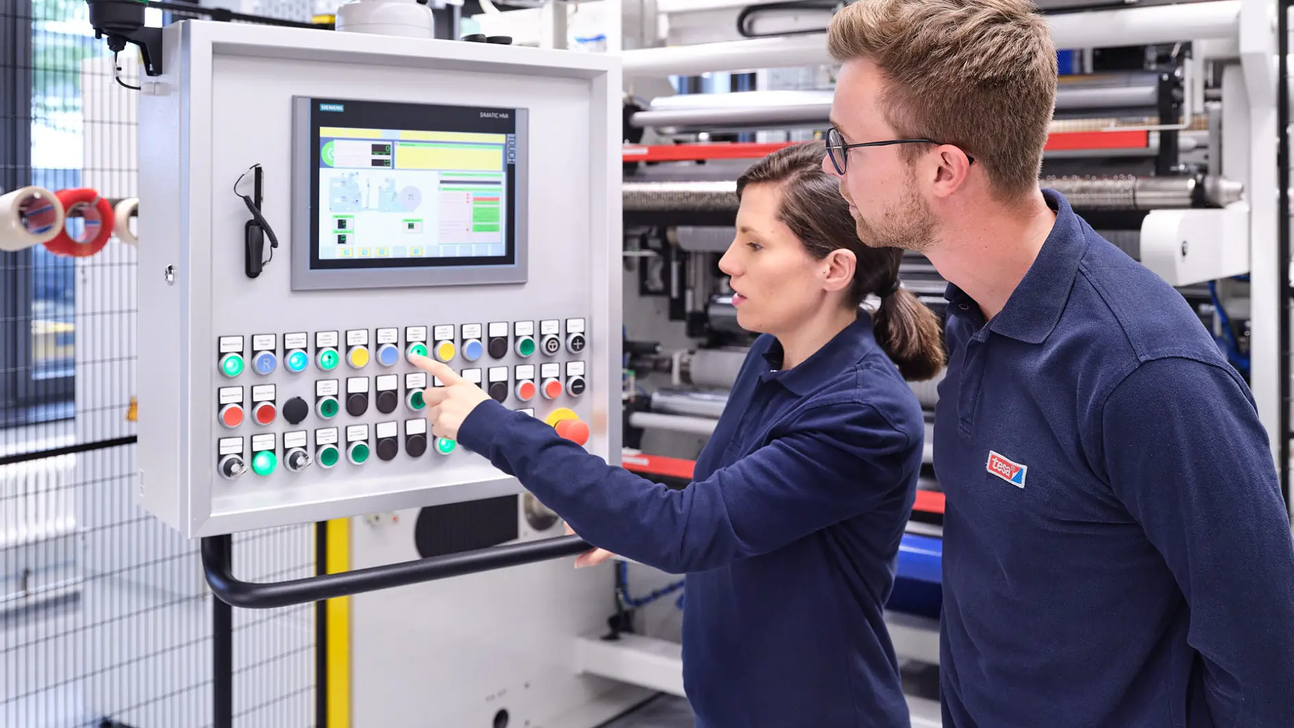 Two people in blue shirts are standing in front of a control panel with multiple buttons and a digital display. One person is pointing at the screen, while the other observes. tesa tape is used throughout the machinery visible in the background.
