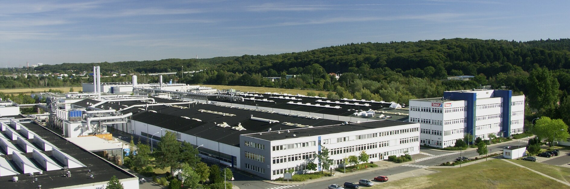 Aerial view of an industrial complex with several large, white-roofed buildings surrounded by greenery. A few trucks are parked on a paved area. In the background, there is a forested area under a clear blue sky. (This text has been generated by AI)