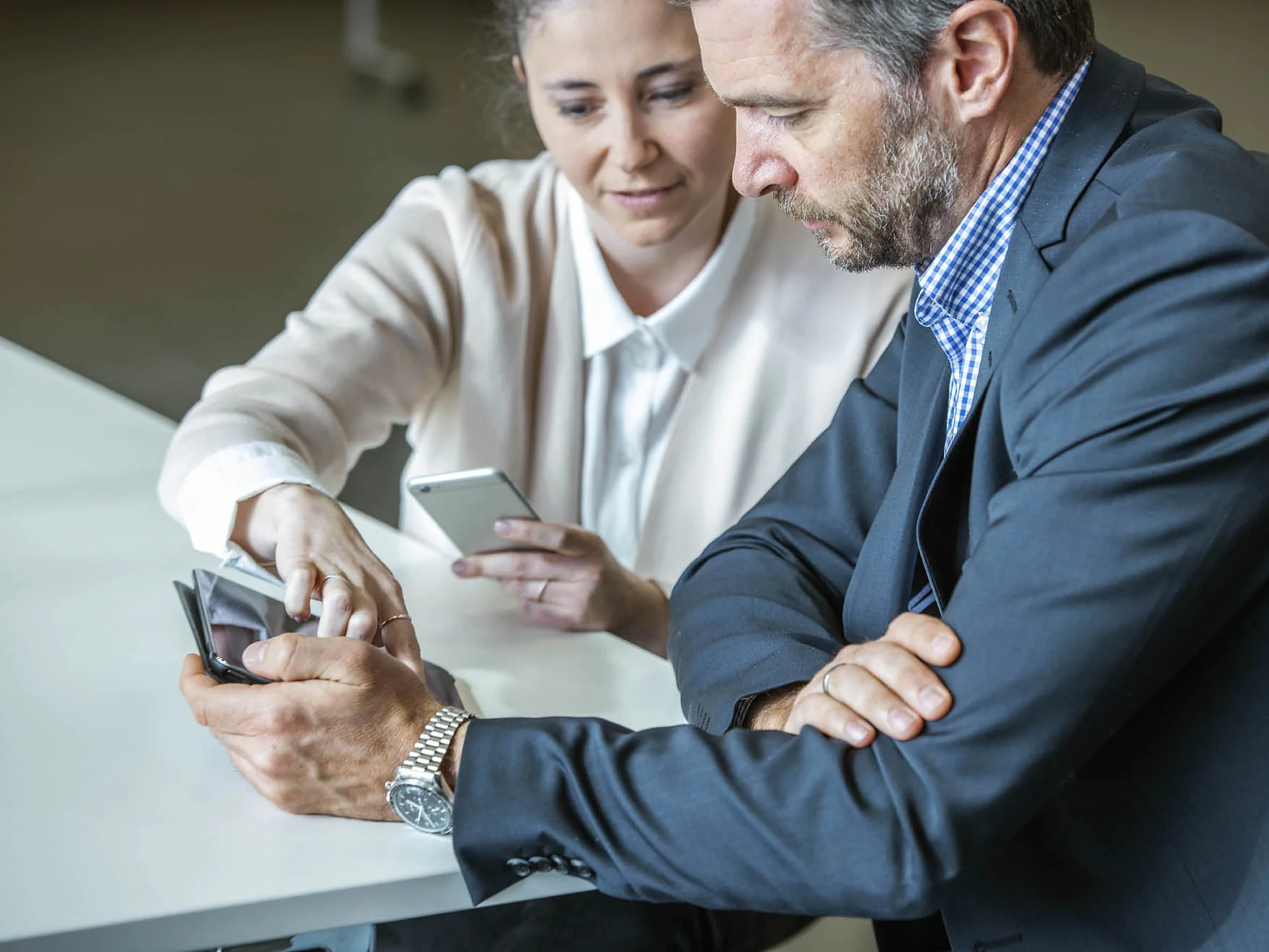 A man and a woman sit at a table, both holding smartphones. The woman points at the mans phone, while the man looks on. They appear to be discussing something on the phone. Both are dressed in business attire. (This text has been generated by AI)