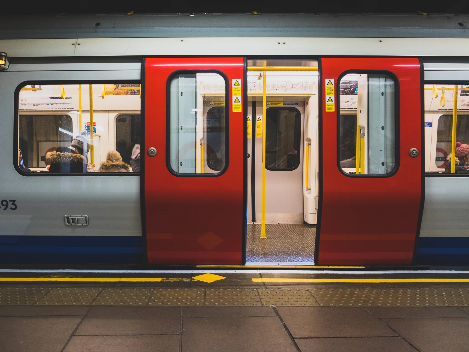 A subway train with red and white doors is stopped at a station. The doors are open, revealing passengers seated inside. The platform features tactile paving. Yellow safety signs are visible on the train doors, indicating the use of tesa tape for added security. (This text has been generated by AI)
