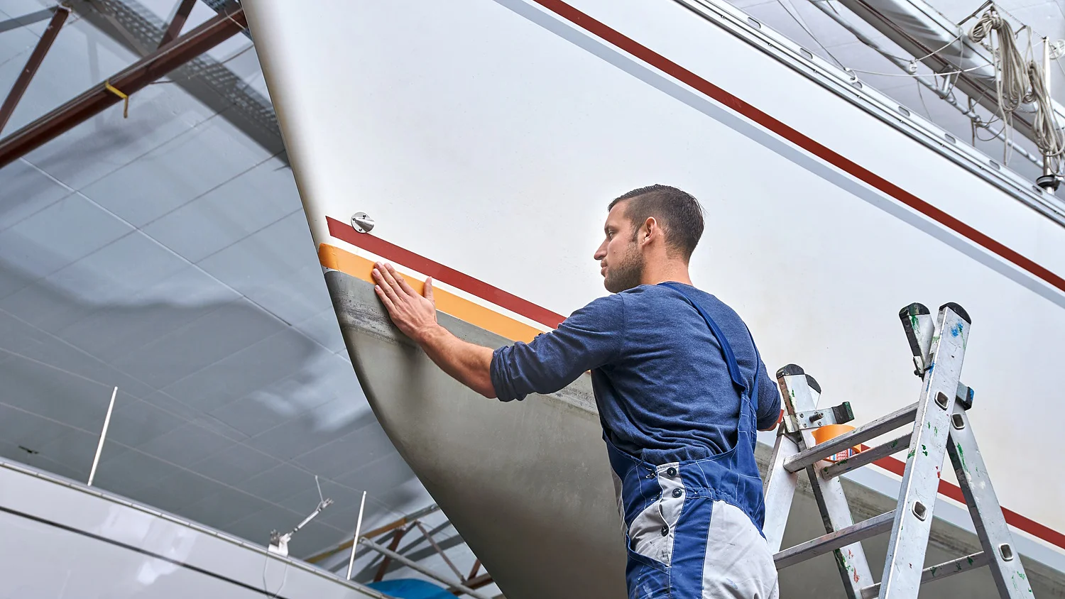 A man in blue overalls stands on a ladder beside a large white sailboat in a workshop. He is examining the boats hull, which features a decorative red and yellow stripe created using tesa tape. The workshop ceiling is visible above. (This text has been generated by AI)