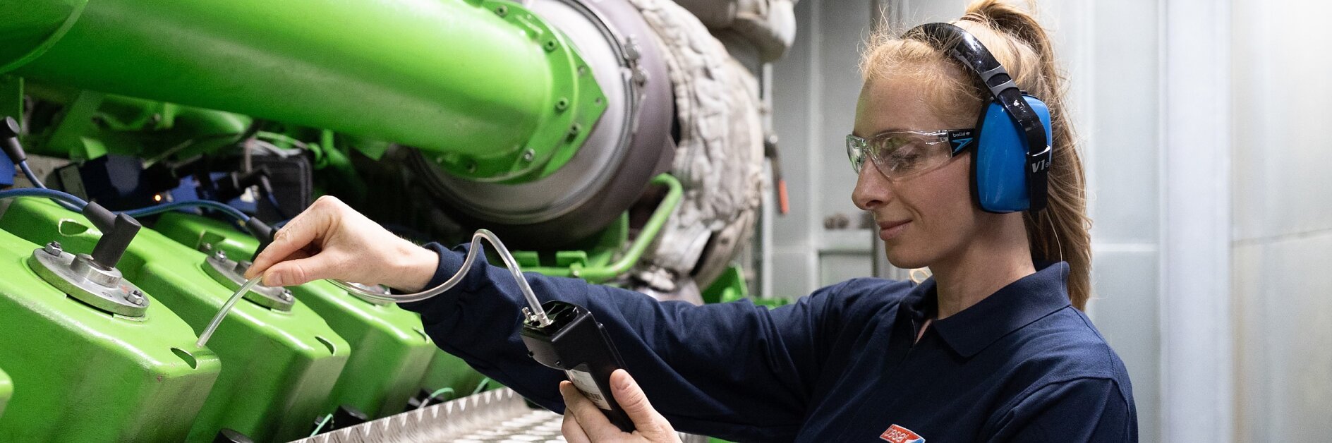 A person wearing safety goggles and earmuffs inspects a large industrial machine with green pipes. They hold a tool in one hand and a tesa tape device in the other, focusing on the machinerys details. The setting is a well-lit industrial area. (This text has been generated by AI)