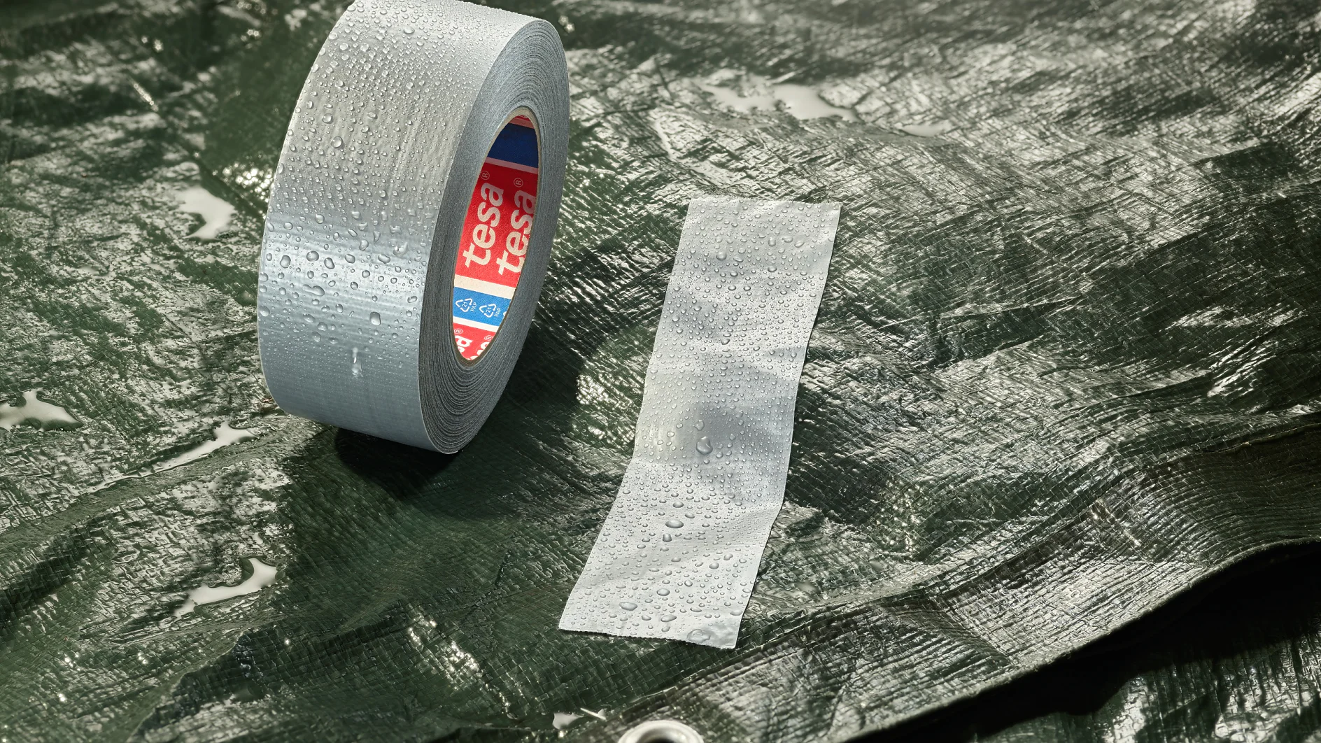 A roll of grey adhesive tesa tape and a strip of tesa tape are placed on a wet, dark green tarpaulin. The tarpaulin has a metal eyelet, and water droplets are visible on the surface. (This text has been generated by AI)