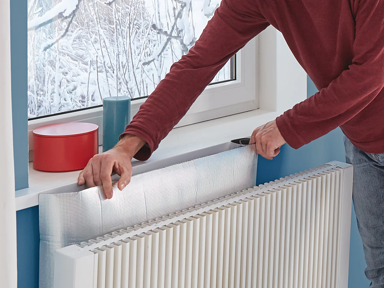 A man in a red sweater and jeans installs foil insulation behind a radiator using tesa tape. A snowy scene is visible outside the window, and the room has blue walls with white curtains. (This text has been generated by AI)