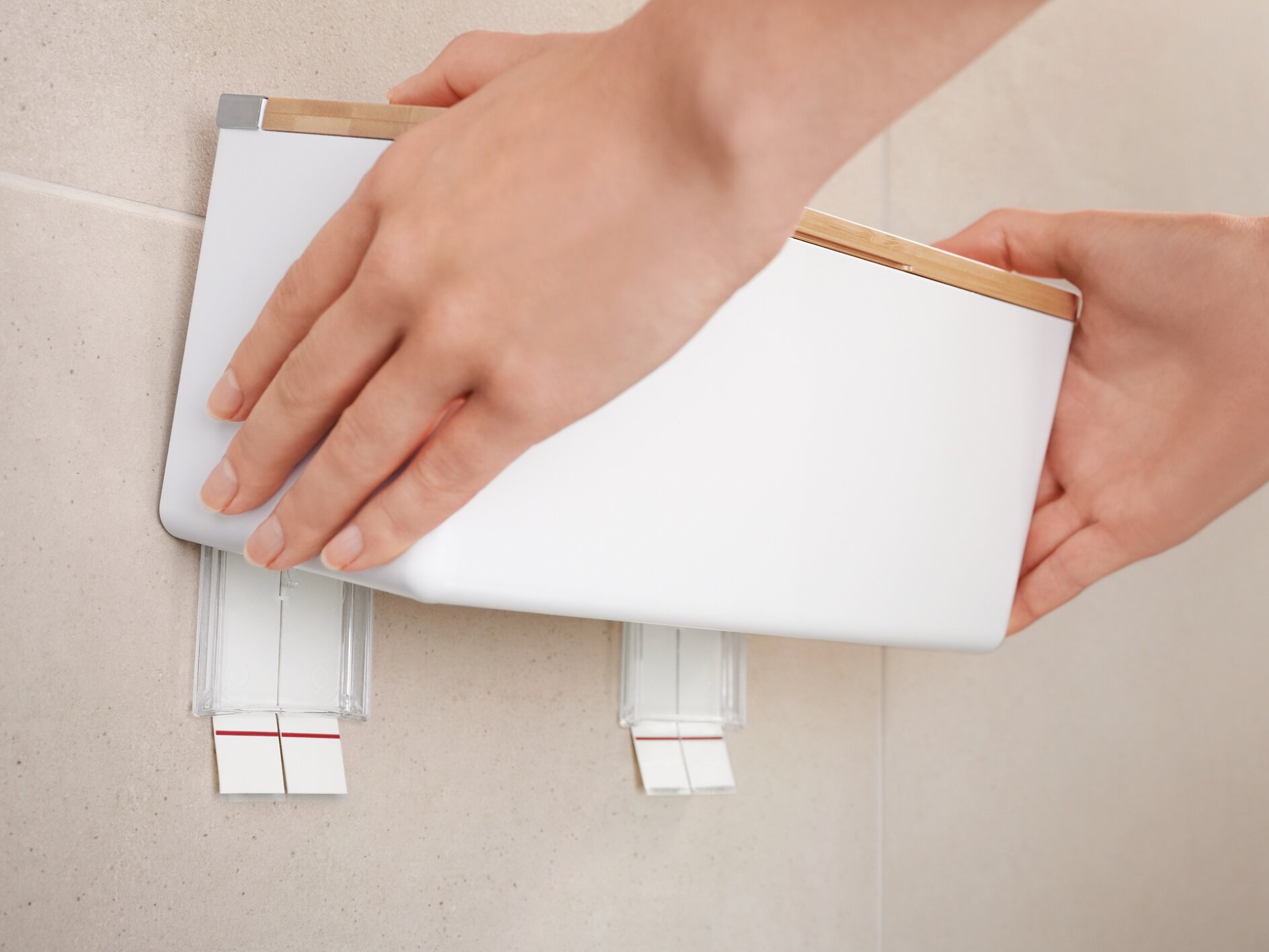 A person is mounting a white, rectangular device onto a wall using two tesa tape adhesive strips. The hands are adjusting the device to ensure it is level. The wall is light-colored, and the scene is indoors. (This text has been generated by AI)