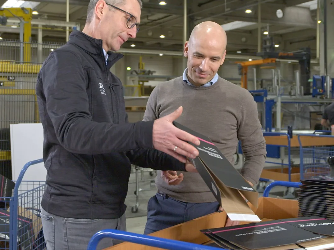 Two men in a factory setting examine printed packaging materials atop a wooden cart. The man on the left, wearing glasses and a black jacket, gestures towards the materials. The man on the right is bald, wearing a grey sweater, and looks attentively at the tesa tape used on them. (This text has been generated by AI)