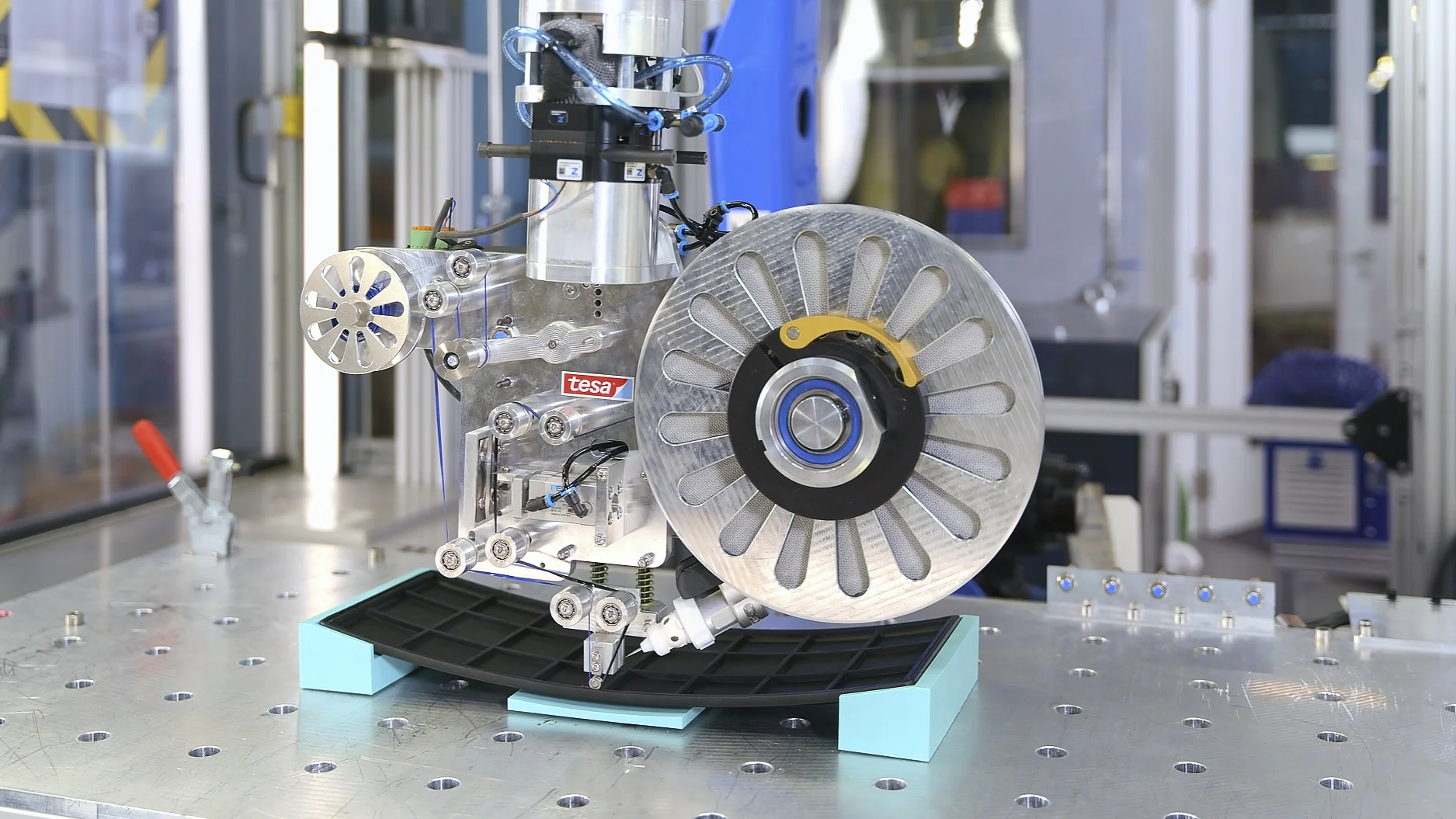 A close-up of a complex mechanical device with exposed gears and wiring, featuring a large metallic disc at the center, situated on a metal worktable in an industrial or laboratory setting. tesa tape is visible among the components, securing wires and parts in place. (This text has been generated by AI)