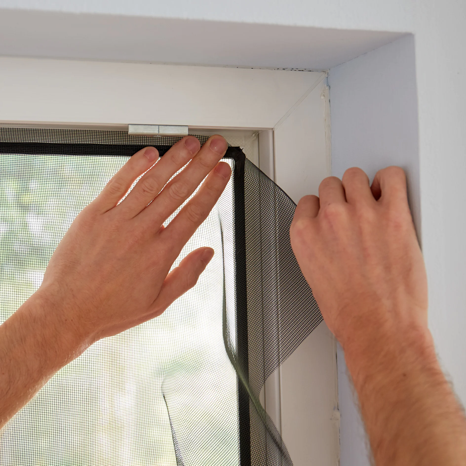 Hands are seen installing a black window screen on a white window frame. One hand holds the screen in place while the other presses it against the frame using tesa tape. Soft outdoor light is visible through the screen. (This text has been generated by AI)