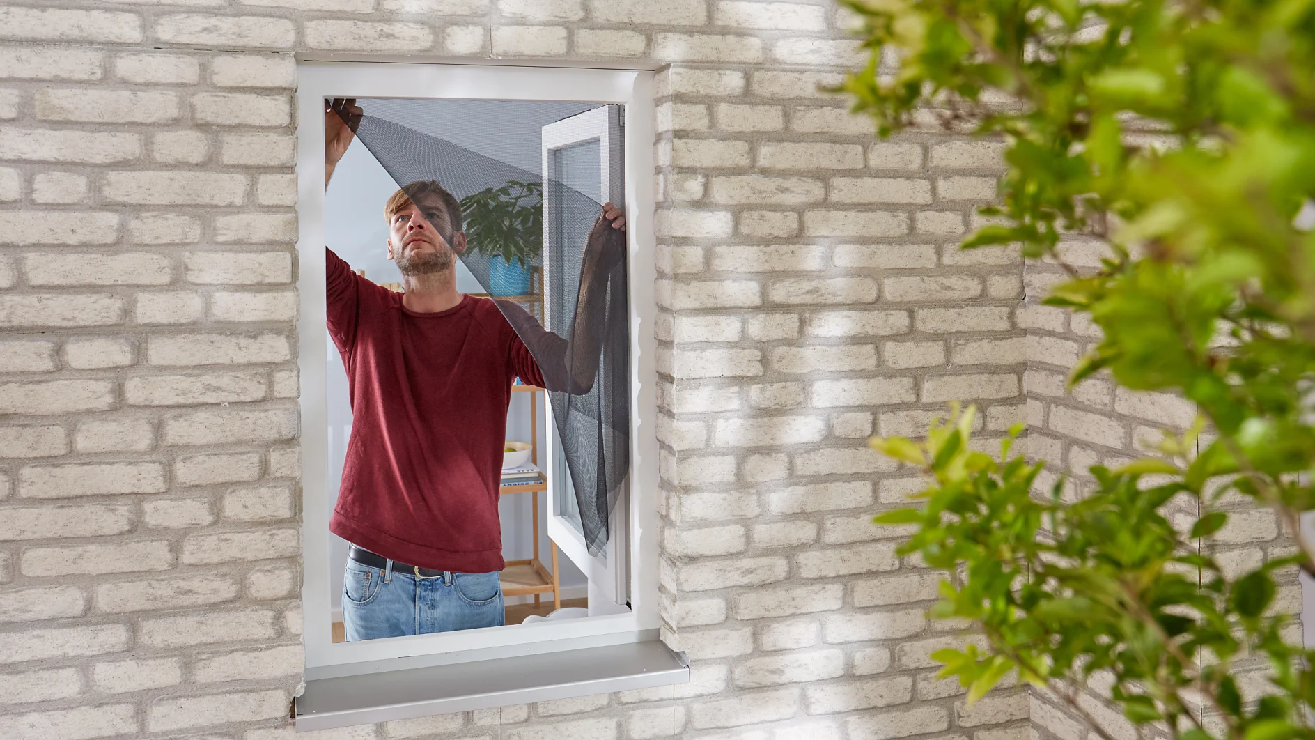 A man in a red shirt and jeans is installing a black mesh screen on a window in a brick wall using tesa tape. The window is open, and there are green leaves partially visible in the foreground. (This text has been generated by AI)
