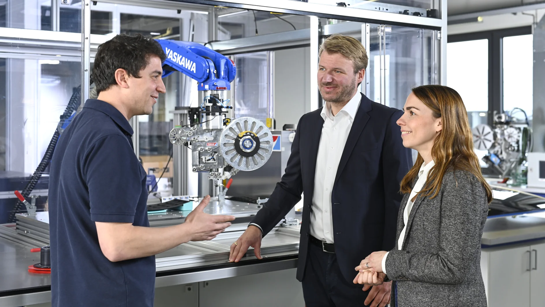 Three people stand in a modern industrial setting, with a blue robotic arm labeled “Yaskawa” and various machinery in the background. One person holds a round machine part wrapped with tesa tape while talking to the other two. (This text has been generated by AI)