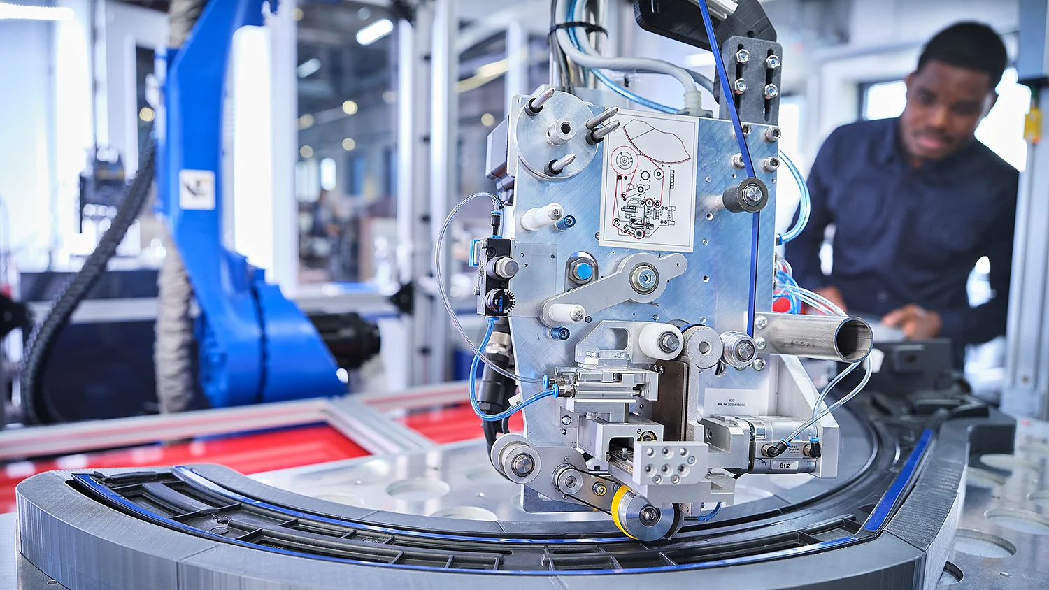 A man stands in the background observing an industrial robotic arm operating on a curved track. The arm is equipped with intricate machinery and wires, set in a modern manufacturing environment with various equipment, including tesa tape, visible. (This text has been generated by AI)