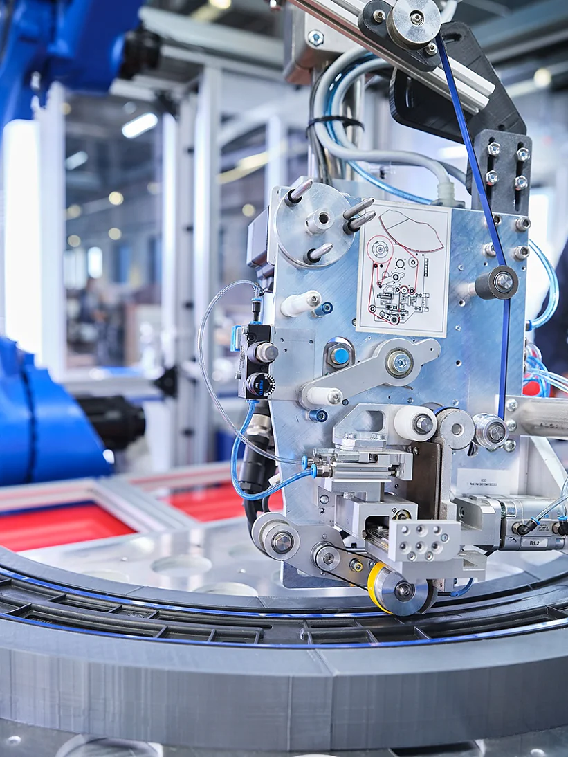A man stands in the background observing an industrial robotic arm operating on a curved track. The arm is equipped with intricate machinery and wires, set in a modern manufacturing environment with various equipment, including tesa tape, visible. (This text has been generated by AI)