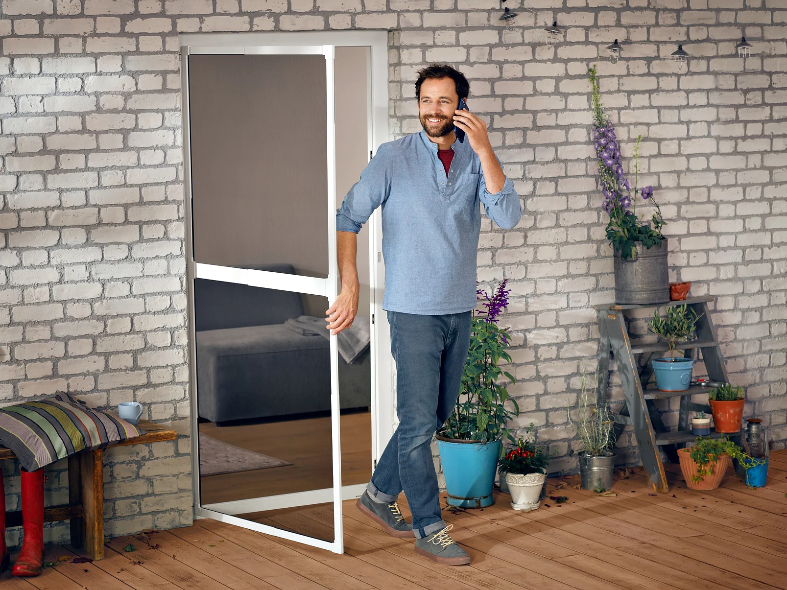 A man stands at the entrance of a patio, talking on a phone. Hes on a wooden deck next to a brick wall with a window and potted plants. A pair of red boots can be seen under a bench with a pillow. Strings of lights hang above, secured with tesa tape. (This text has been generated by AI)