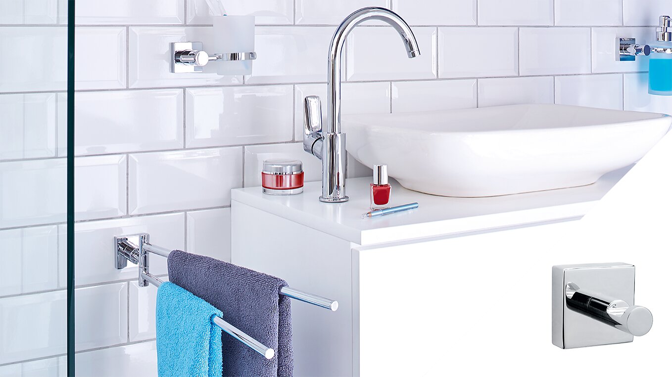 A modern bathroom counter with a white rectangular sink, chrome faucet, and accessories including red nail polish and cream jars. A blue towel hangs on a chrome bar. White subway tiles create the wall backdrop. A square hook is inset in the corner using tesa tape. (This text has been generated by AI)