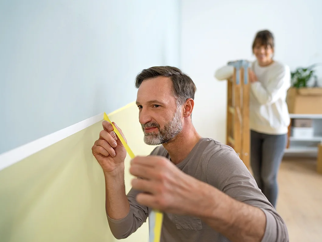 A man is carefully measuring a section of a light green wall with yellow tesa tape. Behind him, a woman is carrying a wooden step ladder. The room contains boxes and a shelf with a plant, suggesting a home improvement project. (This text has been generated by AI)