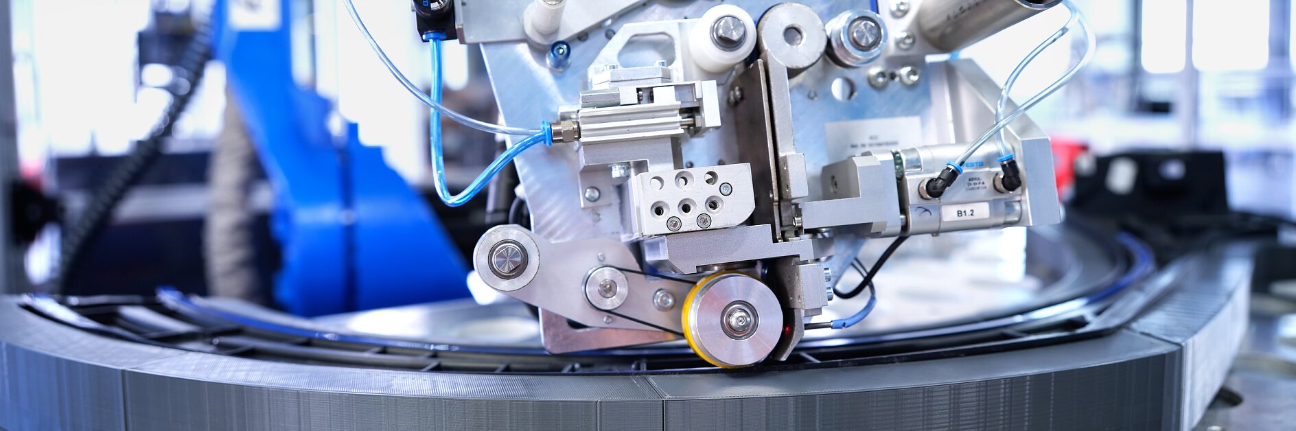 Close-up of industrial machinery with various interconnected metal components, wires, and tubes. The apparatus appears to be operating on a circular track or component. There are pieces of tesa tape visible on some sections, suggesting their use in securing parts or insulating connections. The background shows a blurred interior of a manufacturing facility. (This text has been generated by AI)