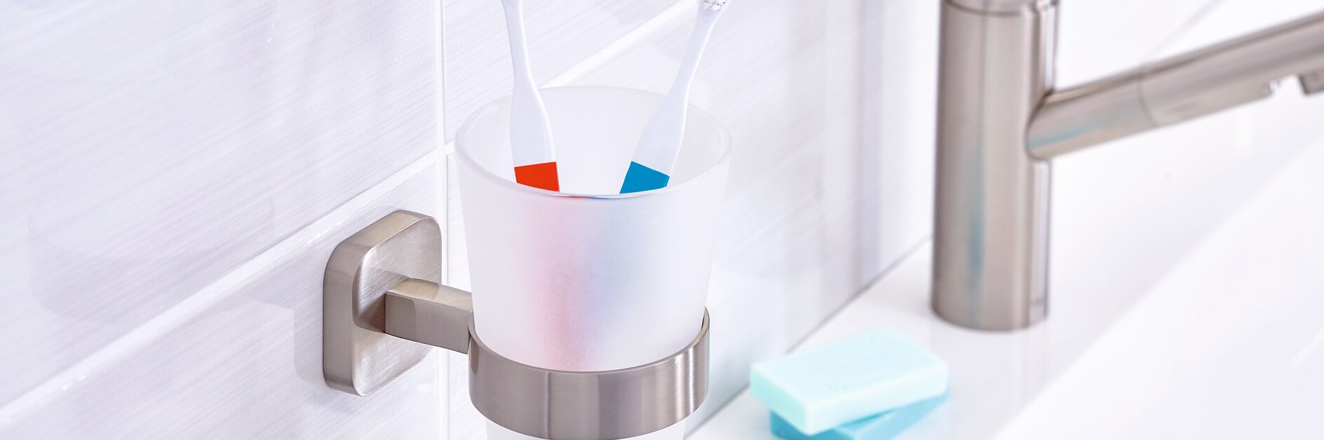 A bathroom scene featuring a matte white toothbrush holder attached to a wall with tesa tape, holding two toothbrushes with blue and red stripes. A silver faucet is on the right, along with a bar of soap on a white countertop. (This text has been generated by AI)