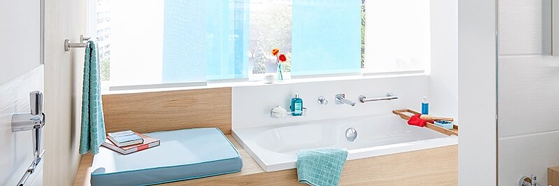 A modern bathroom features a wooden-accented bathtub next to a window with blue curtains. A bath tray holds a red book and blue towel. Toiletries and a potted plant are arranged neatly on the shelf. The space is bright and well-organized, with tesa tape ensuring seamless finishes throughout the room. (This text has been generated by AI)