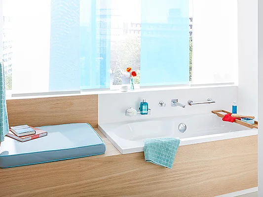 A modern bathroom features a wooden-accented bathtub next to a window with blue curtains. A bath tray holds a red book and blue towel. Toiletries and a potted plant are arranged neatly on the shelf. The space is bright and well-organized, with tesa tape ensuring seamless finishes throughout the room. (This text has been generated by AI)
