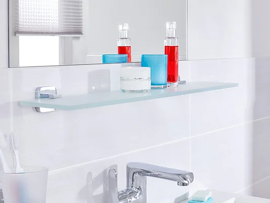 A modern bathroom showcases a glass shelf with blue and white candles alongside two red bottles. Below the shelf, a sink with a chrome faucet is partially visible. Toothbrushes in a holder are positioned on the left, while a hairdryer is mounted on the right using tesa tape. (This text has been generated by AI)