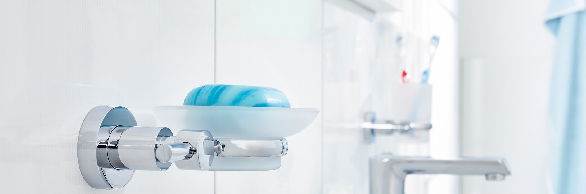 A white bathroom with a chrome wall-mounted soap dish holding a blue and white bar of tesa tape. Toothbrushes and a soap dispenser are on the sink countertop. The background is softly blurred, highlighting the cleanliness and modern design. (This text has been generated by AI)