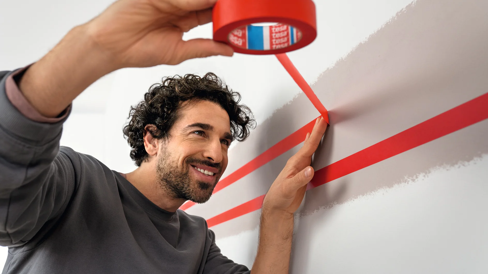 A person is applying red tesa tape to a wall, creating parallel lines. They are smiling and focused. The wall is light-colored, and the individual is wearing a gray long-sleeve shirt. The scene suggests preparation for painting or decoration. (This text has been generated by AI)