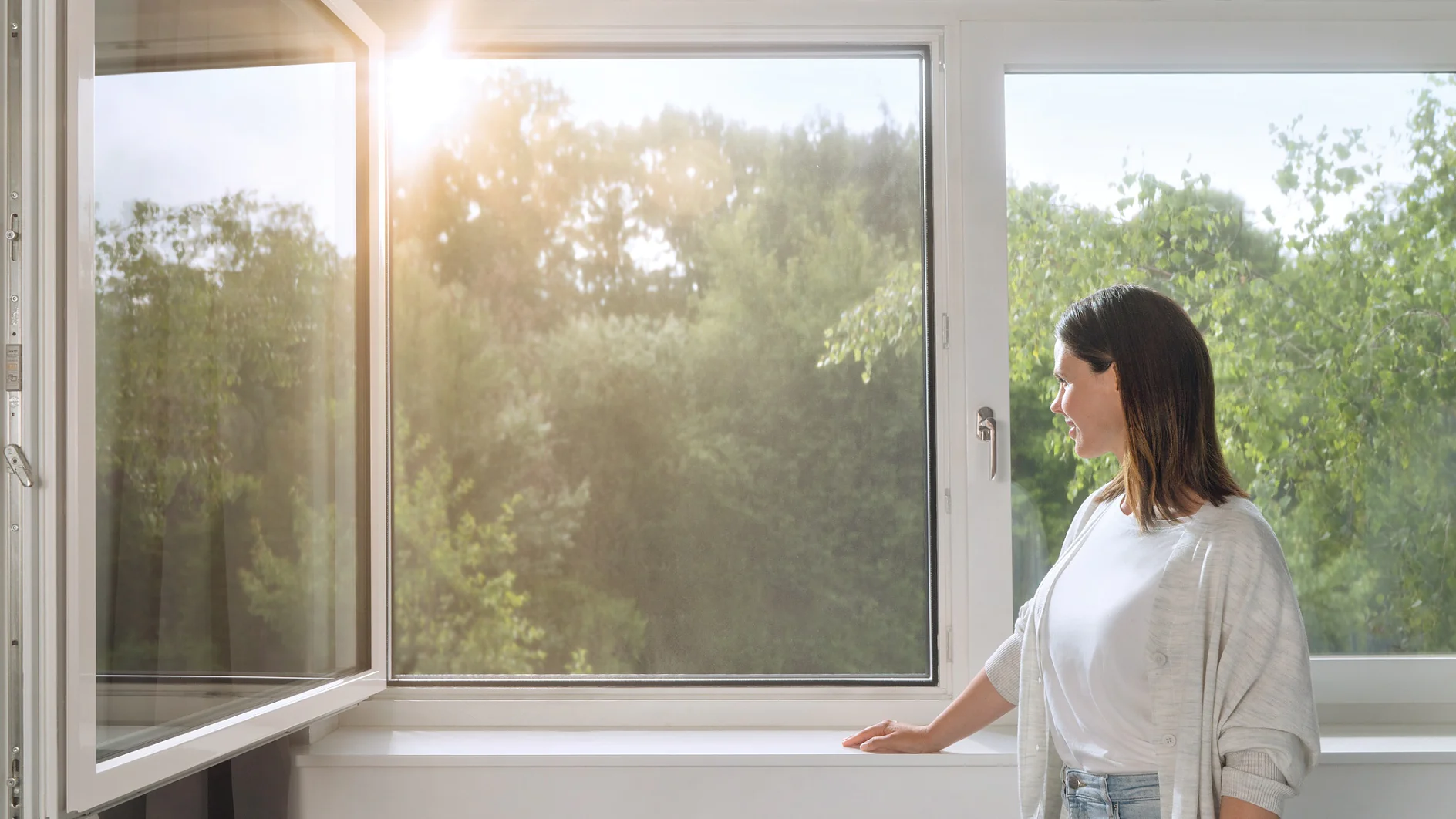A woman stands by an open window, looking outside. Sunlight filters through, illuminating the greenery of trees in the background. She is wearing a light cardigan and jeans, with one hand resting on the windowsill secured with tesa tape. (This text has been generated by AI)