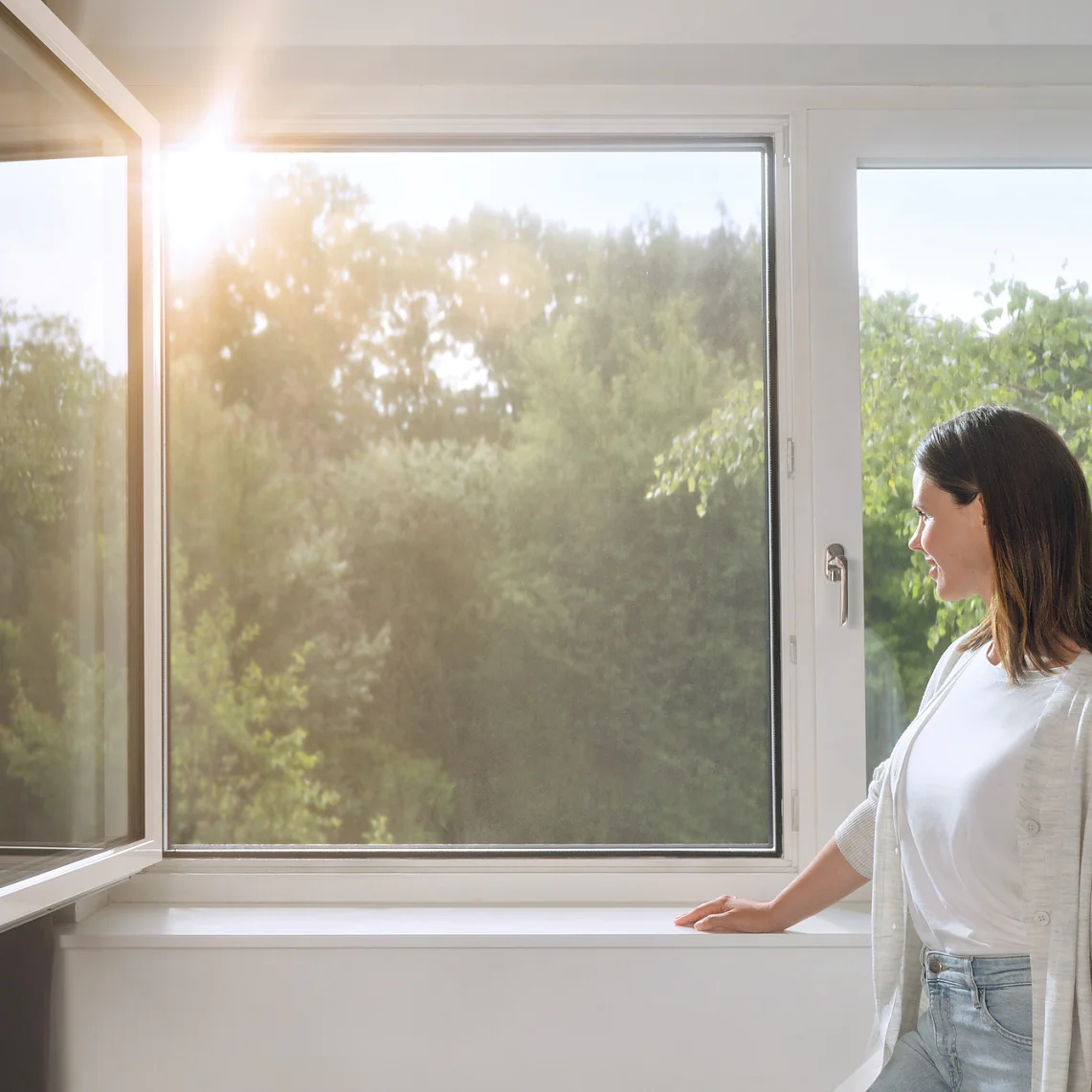 A woman stands by an open window, looking outside. Sunlight filters through, illuminating the greenery of trees in the background. She is wearing a light cardigan and jeans, with one hand resting on the windowsill secured with tesa tape. (This text has been generated by AI)