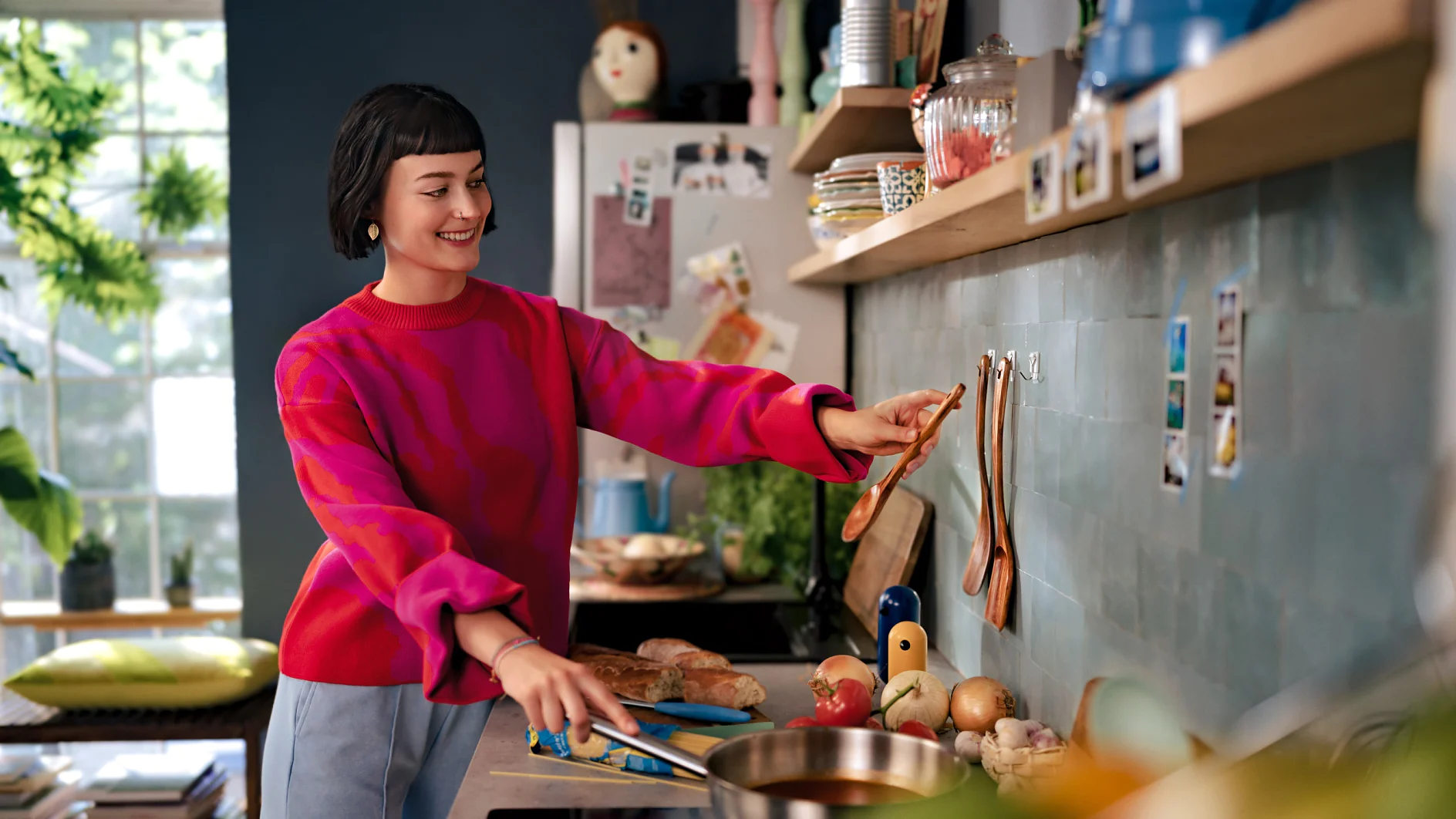 A woman in a red sweater arranges wooden spoons on a wall in a kitchen using tesa tape. She stands beside a stove with a pan, bread, and vegetables. Shelves holding jars and a plant are visible, with a large window providing natural light in the background. (This text has been generated by AI)
