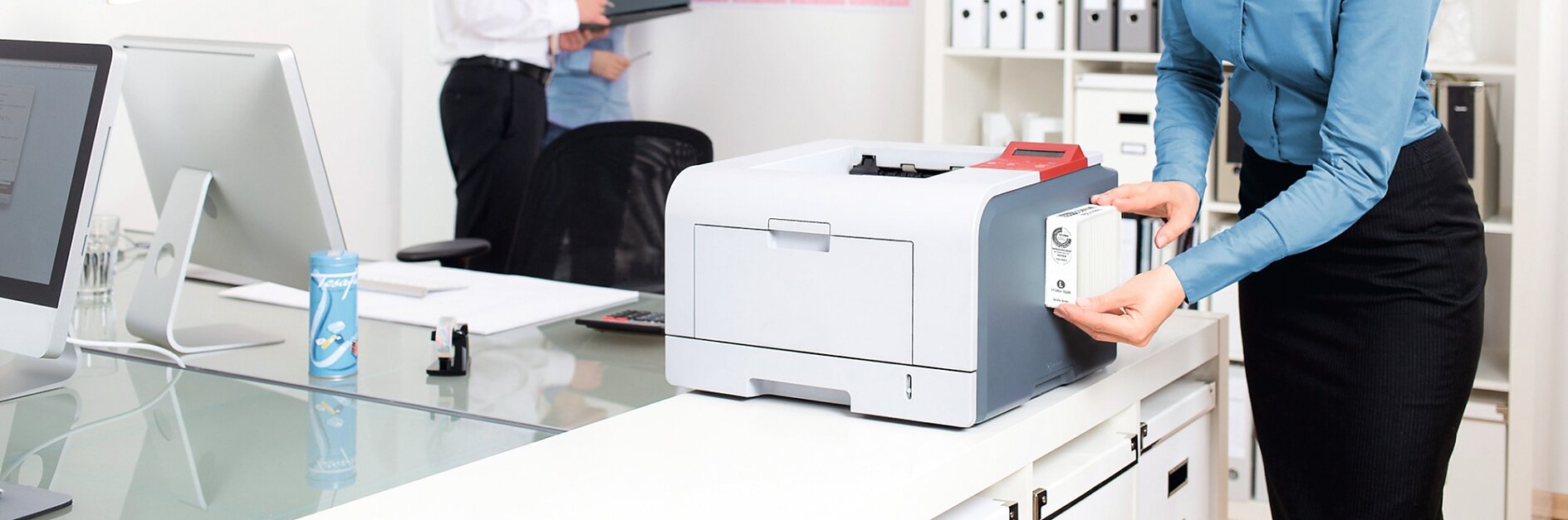 A woman in a blue shirt replaces the paper in a printer in an office. In the background, a man and a woman stand near a wall calendar, discussing. The office is bright, with a desk, lamp, cup, tesa tape dispenser, and clock visible. (This text has been generated by AI)