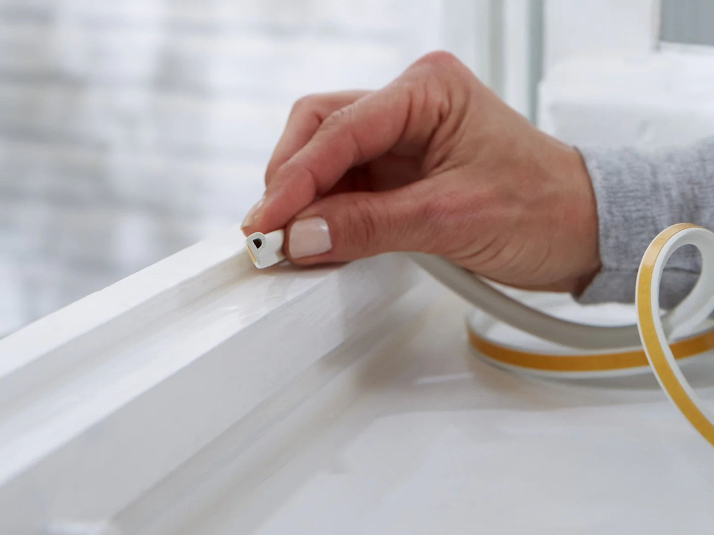 A hand is placing tesa tape weather stripping onto a white window frame. The background shows a blurred exterior wall. The weather stripping is beige with a peel-off backing, suggesting a home improvement or insulation task. (This text has been generated by AI)