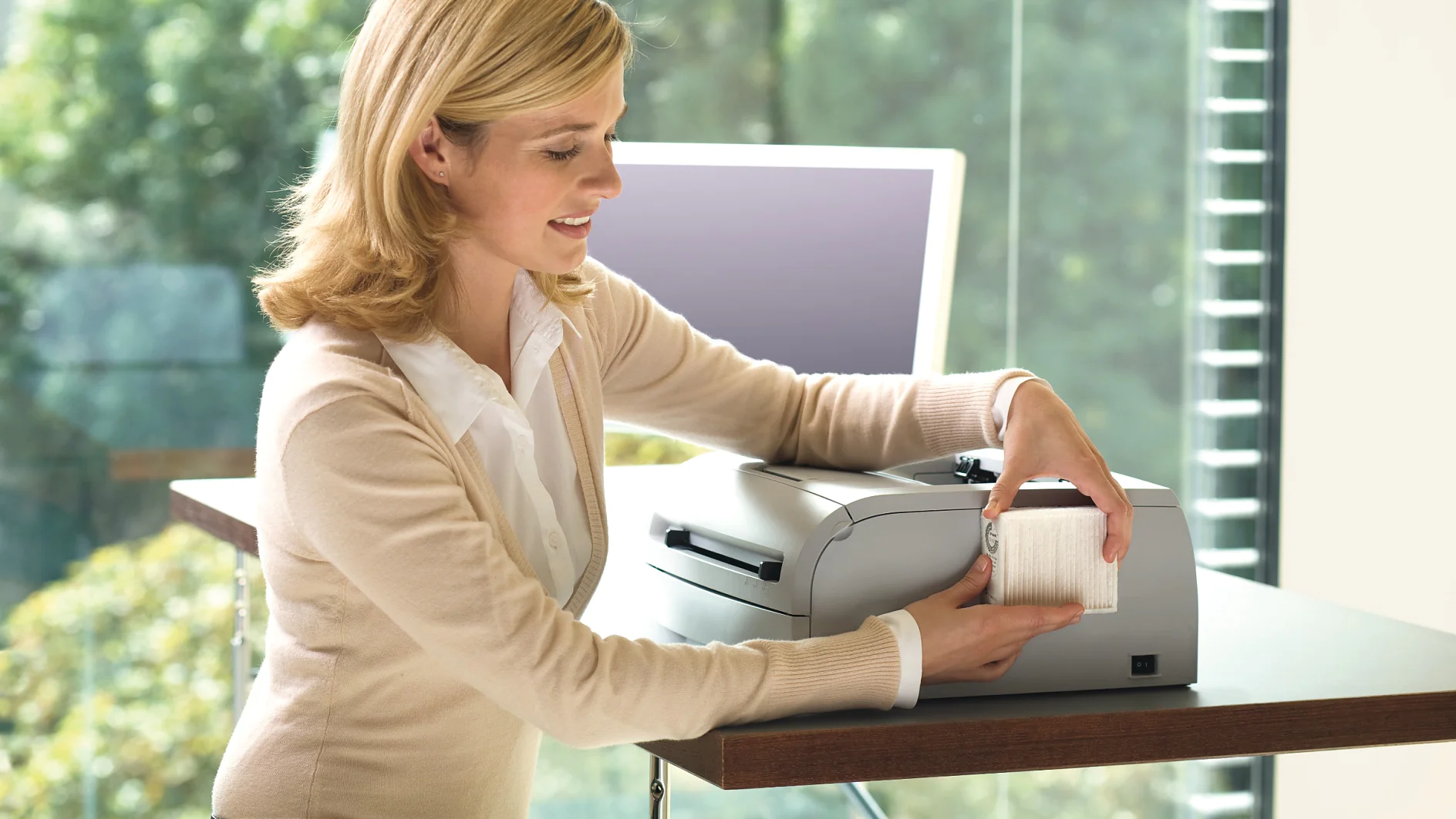 A person with blonde hair is seated at a desk, using a printer. They are smiling and removing a printed sheet from the device, with tesa tape on hand for any quick fixes needed. There is a computer monitor in the background. Sunlight streams through large windows with a view of green trees. (This text has been generated by AI)